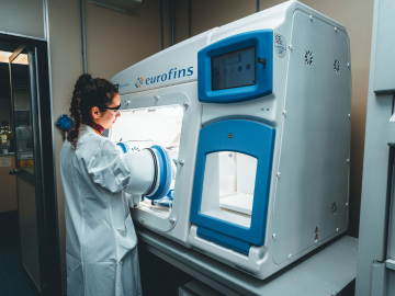 Una científica con bata y gafas protectoras trabaja en un laboratorio. Utiliza una máquina avanzada para realizar pruebas microbiológicas estériles. El entorno es limpio y profesional, lo que pone de relieve la importancia de la precisión y la seguridad en la investigación científica.