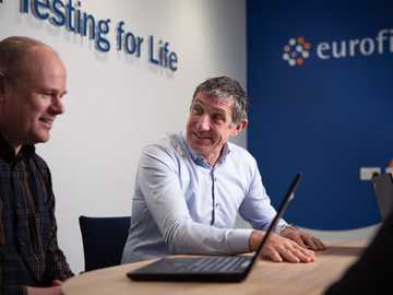 Dos consultores conversan en una oficina moderna, con el logotipo de Eurofins visible al fondo. La imagen capta un entorno empresarial de colaboración centrado en los productos sanitarios