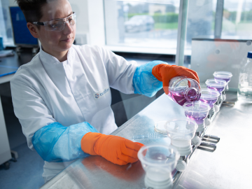 Una técnica de laboratorio con bata, guantes y gafas protectoras, realizando análisis de medios de cultivo y bioindicadores en un laboratorio de Eurofins BioPharma Product Testing. La técnica evalúa la calidad y efectividad de los medios de cultivo utilizados para el crecimiento de microorganismos, asegurando la fiabilidad de los bioindicadores en las pruebas microbiológicas y el control de calidad de productos farmacéuticos.
