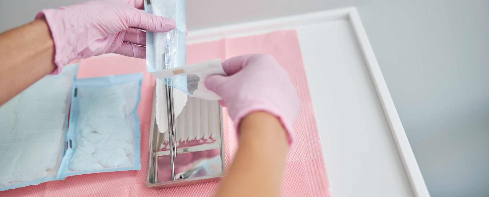 Two gloved hands are opening a plastic package containing a scalpel on a white table with a pink protective sheet