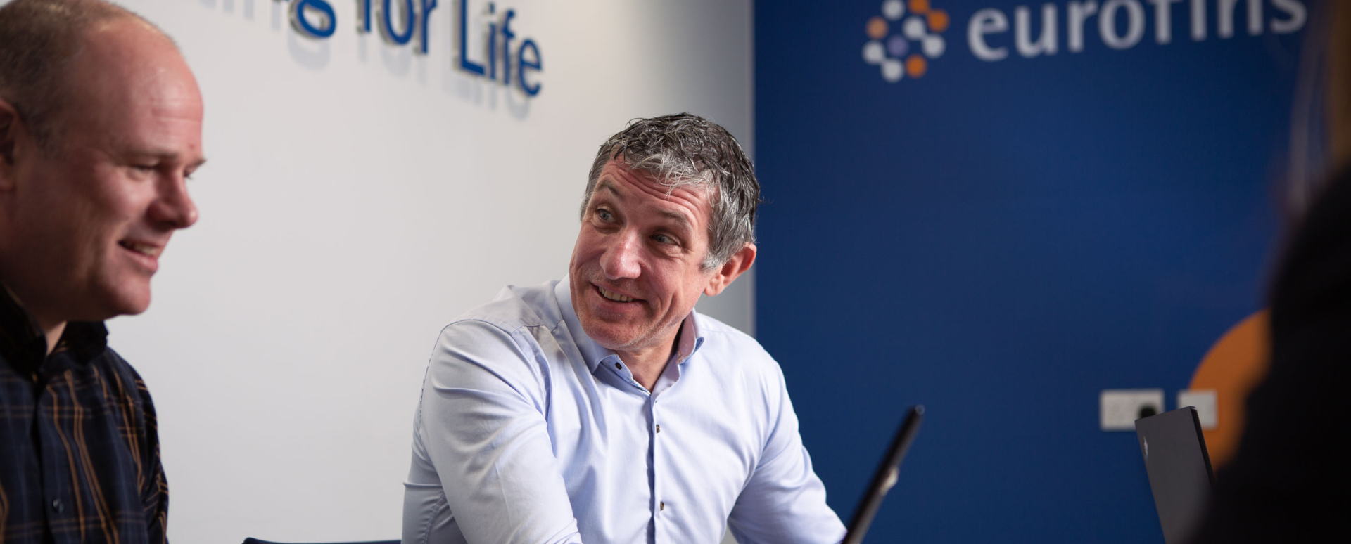 Dos consultores conversan en una oficina moderna, con el logotipo de Eurofins visible al fondo. La imagen capta un entorno empresarial de colaboración centrado en los productos sanitarios