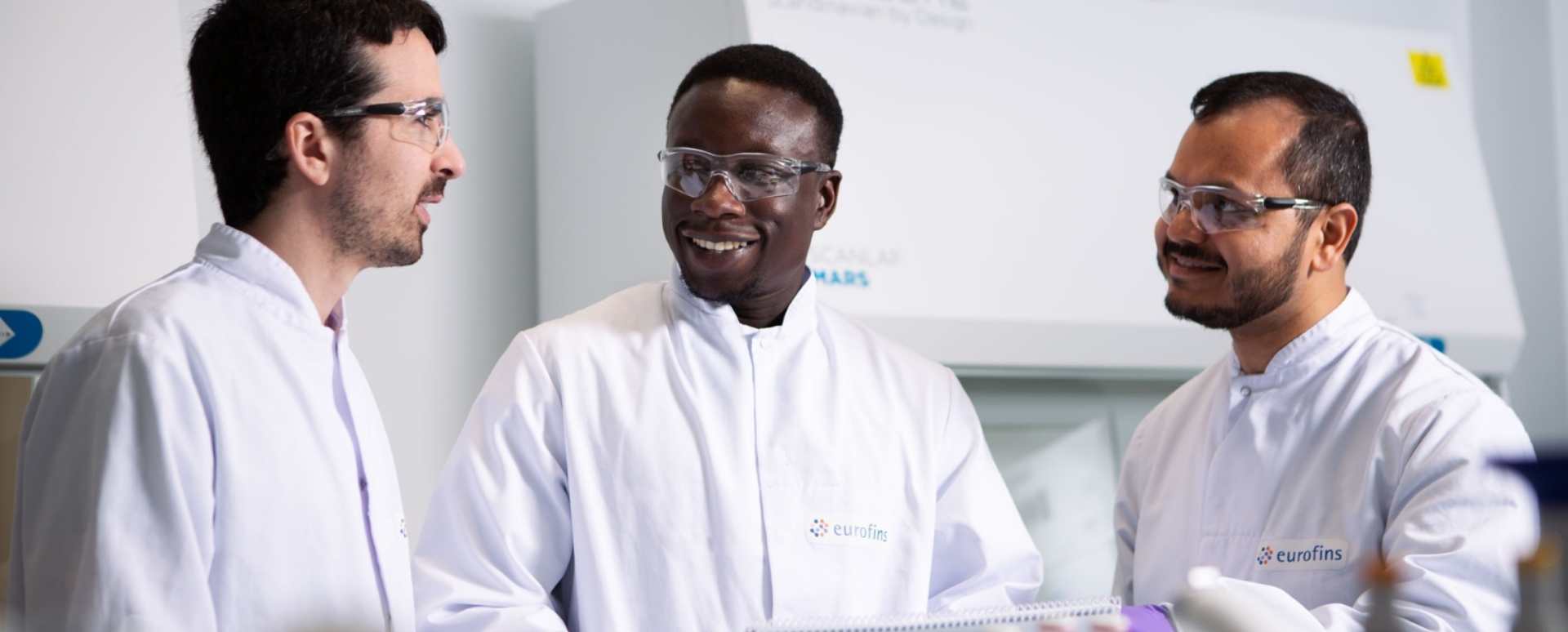 Tres asesores con batas blancas de laboratorio y gafas de seguridad hablando.