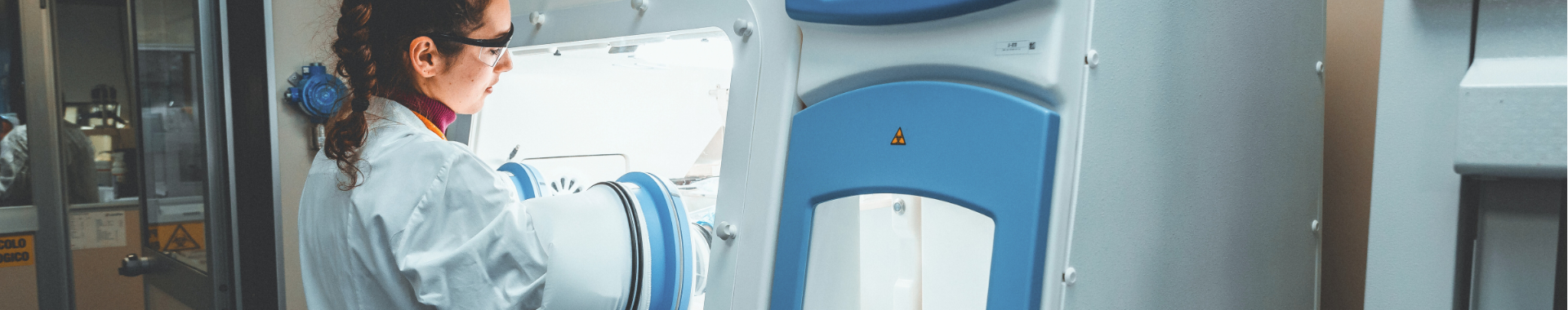 Una científica con bata y gafas protectoras trabaja en un laboratorio. Utiliza una máquina avanzada para realizar pruebas microbiológicas estériles. El entorno es limpio y profesional, lo que pone de relieve la importancia de la precisión y la seguridad en la investigación científica.