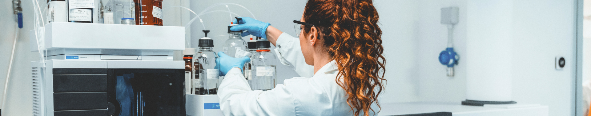 Una científica con bata y gafas protectoras trabaja en un laboratorio. Utiliza una máquina avanzada para realizar análisis de productos no obligatoriamente estériles. El entorno es limpio y profesional, lo que pone de relieve la importancia de la precisión y la seguridad en la investigación científica.