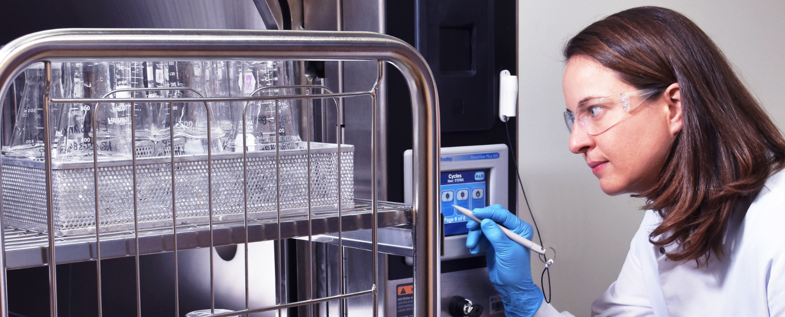 A woman in a lab cost is holding a stylus in her gloved hand to adjust settings on an autoclabe being filled with two shelves of clear beakers in bins.