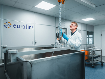 Una científica con bata de laboratorio blanca, gafas de protección y guantes azules realizando pruebas de integridad del cierre del envase, mostrando equipos avanzados y experiencia en pruebas de productos biofarmacéuticos dentro de un entorno de investigación de alta precisión.