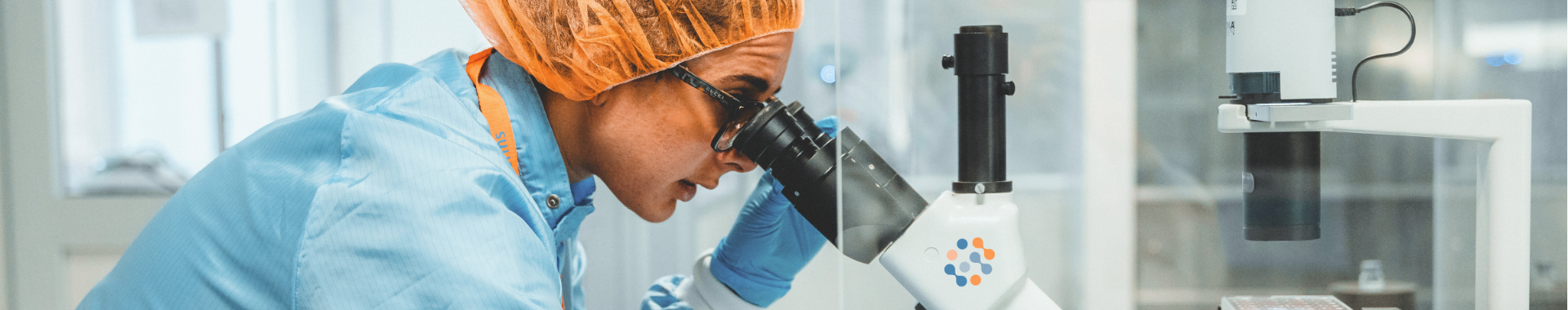 Una técnica de laboratorio con bata, guantes y cofia para el cabello, realizando un análisis al microscopio para la identificación de microorganismos en un laboratorio de Eurofins BioPharma Product Testing. Utilizando tecnologías avanzadas, la técnica trabaja en la identificación de bacterias, mohos y levaduras, proporcionando análisis expertos para detectar contaminantes en productos terminados y rastrear la fuente de la contaminación mediante tipado genético de los microorganismos.