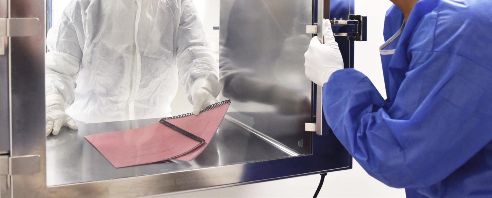 A person in white coverall with white gloves and laboratory safety goggles is handing a bound report with a red cover through a window with a door to a woman with white gloves, blue coverall and a hair net