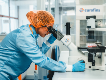 Una técnica de laboratorio con bata, guantes y cofia para el cabello, realizando un análisis al microscopio para la identificación de microorganismos en un laboratorio de Eurofins BioPharma Product Testing. Utilizando tecnologías avanzadas, la técnica trabaja en la identificación de bacterias, mohos y levaduras, proporcionando análisis expertos para detectar contaminantes en productos terminados y rastrear la fuente de la contaminación mediante tipado genético de los microorganismos.