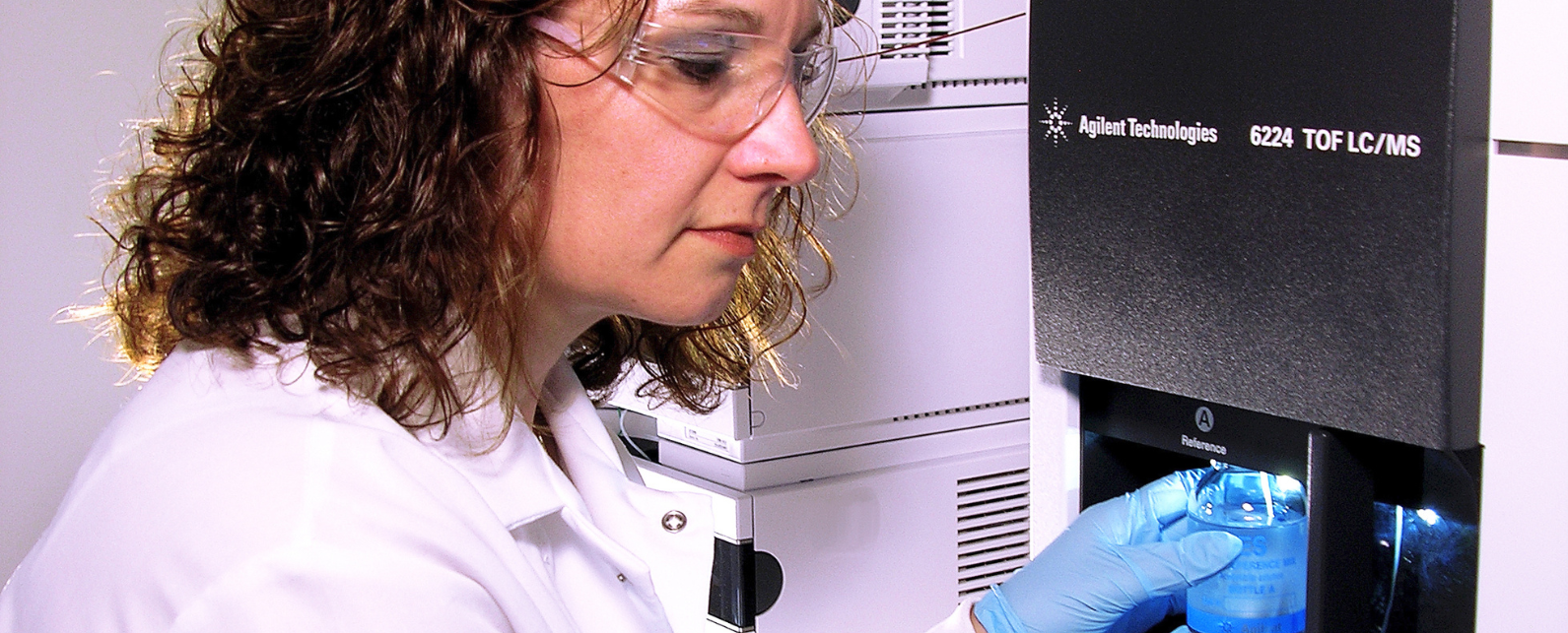 Una mujer morena con bata de laboratorio y gafas sostiene una muestra en un Agilent 6224 TOF LC/MS con las manos enguantadas.