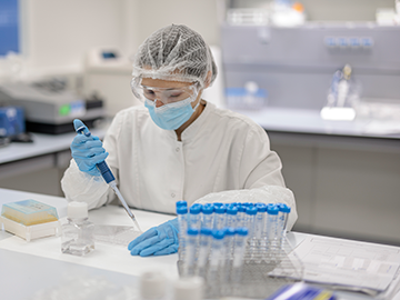 Un investigador en un laboratorio, con bata blanca, guantes, mascarilla y redecilla protectora, utiliza una pipeta para transferir líquido a un recipiente. La estación de trabajo tiene tubos de ensayo, herramientas de laboratorio y otros materiales científicos.
