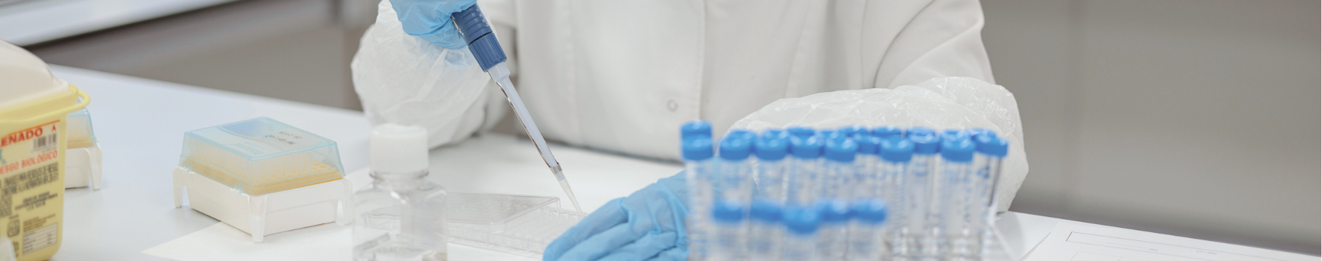 Un investigador en un laboratorio, con bata blanca, guantes, utiliza una pipeta para transferir líquido a un recipiente. La estación de trabajo tiene tubos de ensayo, herramientas de laboratorio y otros materiales científicos.